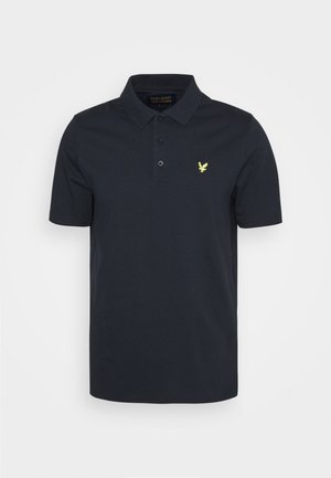 Mand iført sort poloshirt med lille gult logo, lyseblå jeans og hvide sneakers med gennemsigtige grønne såler, der står indendørs.