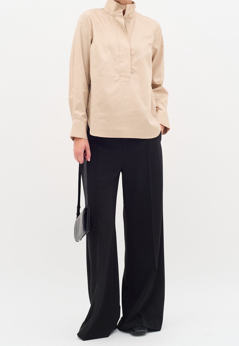 Femme portant un chemisier beige à col haut et manches longues, pantalon noir à jambes larges, tenant un petit sac à main noir à côté d'elle.