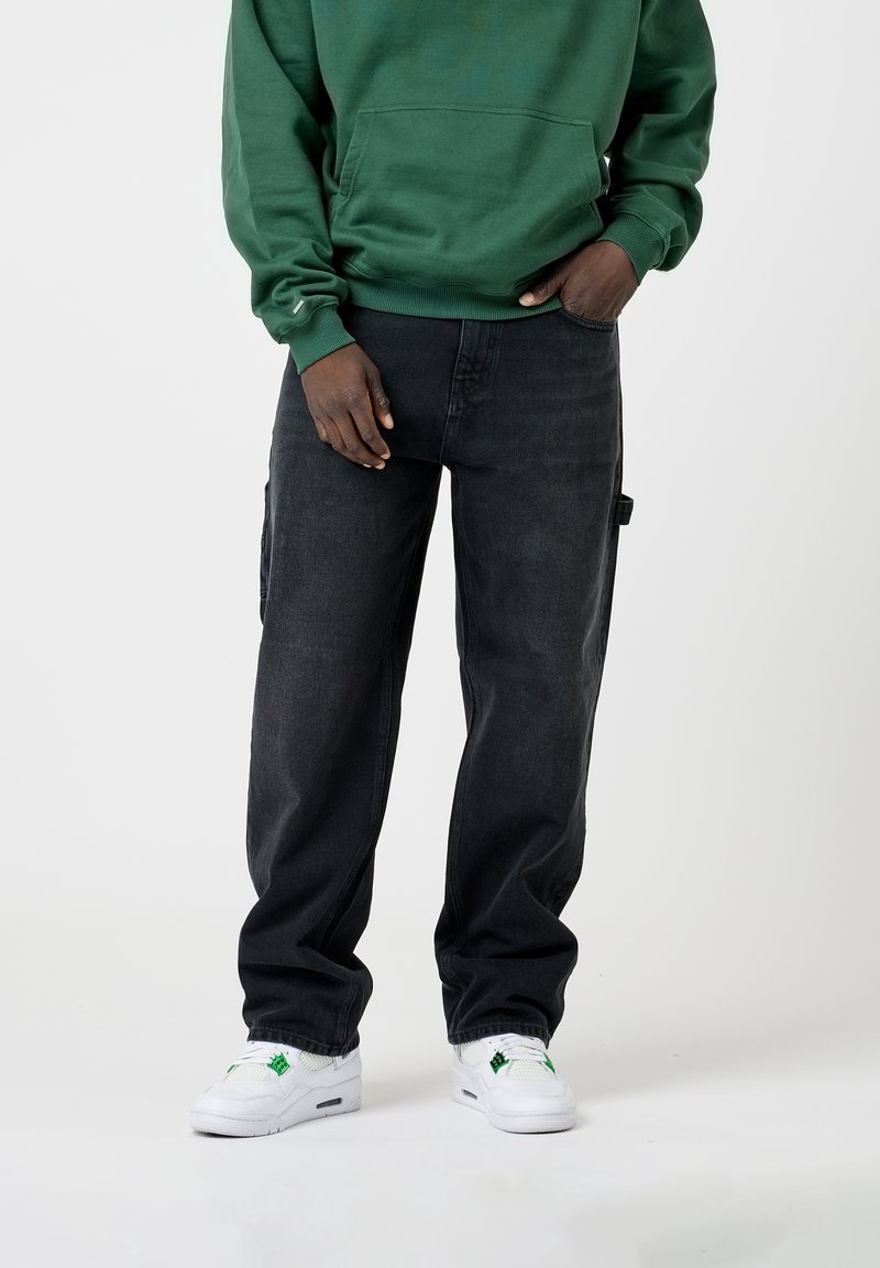 Personne portant un sweat à capuche vert, un jean noir ample et des baskets blanches avec des accents verts, debout avec une main dans la poche devant un fond uni.