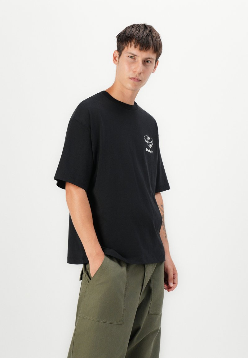 T-shirt nera in cotone con maniche corte, colletto tondo e una piccola grafica bianca sul petto. Indossata con pantaloni verde oliva.