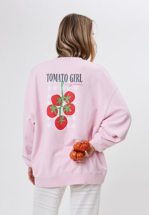 Vrouw met schouderlang haar die een roze sweatvest draagt met de tekst "TOMATO GIRL" en tomaatgrafieken, terwijl ze twee tomaten achter haar rug vasthoudt.