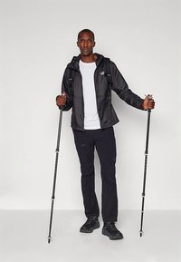 Μαύρο αδιάβροχο μπουφάν με κουκούλα, λευκό μπλουζάκι, μαύρο παντελόνι και μαύρα μποτάκια πεζοπορίας; κρατώντας μ. ορειβατικά μπαστούνια μπροστά σε απλό φόντο.