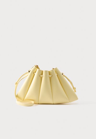Απαλό κίτρινο δερμάτινο τσαντάκι με κορδόνι, κάθετες πτυχές και λεπτό ιμάντα ώμου, τοποθετημένο πάνω σε απλό φωτεινό φόντο.