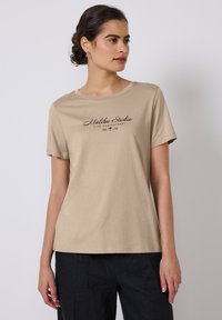 T-shirt en coton beige à col rond et manches courtes. L'avant présente un texte imprimé noir "Malibu Studio" et un accent de palmier.