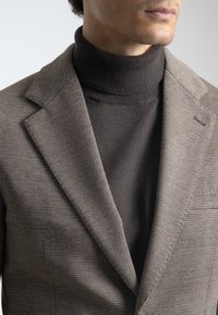 Blazer de cuadros marrón con solapas, superpuesto a un cuello de tortuga marrón oscuro. Tela suave y texturada con finos detalles.