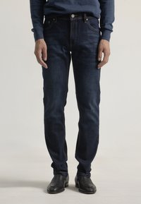 Jeans de denim azul oscuro con corte ajustado, que incluyen tres bolsillos frontales, costuras tonales y cierre de botón. Llevados con zapatos negros.