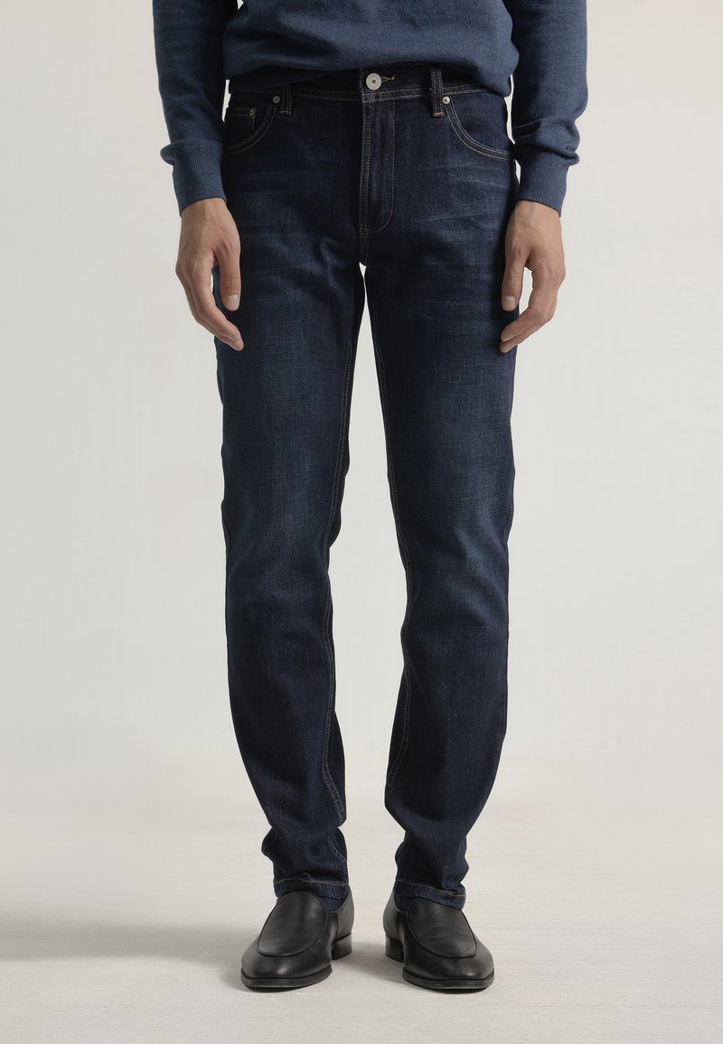 Jeans de denim azul oscuro con corte ajustado, que incluyen tres bolsillos frontales, costuras tonales y cierre de botón. Llevados con zapatos negros.