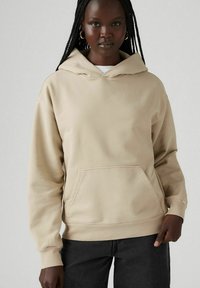 Sudadera capucha beige con un bolsillo delantero, hecha de una tela suave. Presenta un ajuste relajado y puños acanalados, combinada con jeans oscuros.