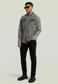 Camicia di denim grigia con chiusura a bottoni e due tasche sul petto, abbinata a jeans skinny neri e stivali neri, su uno sfondo neutro.