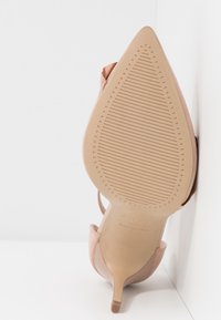 Une seule chaussure à talon haut beige à bout pointu montrant une semelle texturée antidérapante avec un talon aiguille fin sur fond blanc.
