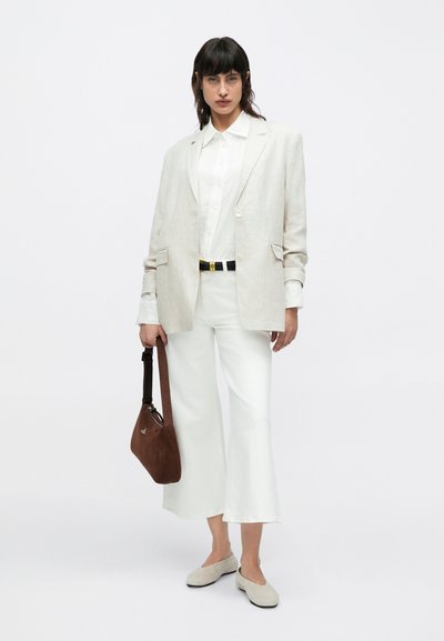 Leinenblazer in Off-White, kombiniert mit einem weißen Hemd und weißen Hosen mit weitem Bein. Braune Wildledertasche und hellgraue Slipper runden den Look ab.