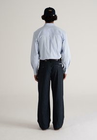 Man in een lichtblauw shirt, zwarte pet, riem en zwarte gestreepte wijde broek, staande met de rug naar de camera tegen een effen achtergrond.
