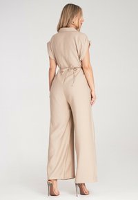 Beżowy kombinezon z krótkimi rękawami zrolowanymi, paskiem w talii, szerokimi nogawkami i kołnierzykiem. Gładka tekstura z minimalnymi detalami.