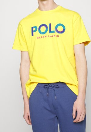 Osoba ubrana w żółtą koszulkę z napisem „POLO RALPH LAUREN” oraz niebieskie spodnie z troczkami na jednolitym jasnym tle.