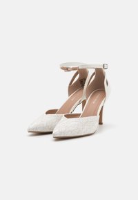 Tacchi bianchi a punta con una texture floreale simile a pizzo, cinturino alla caviglia e tacco sottile. Fodera interna liscia e sottili accenti beige.
