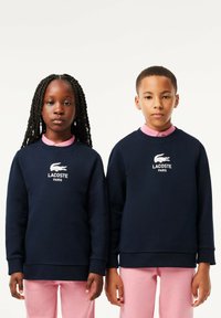 Sweatshirts bleu marine avec des poignets côtelés, arborant le logo blanc de Lacoste et le texte « PARIS » ; portés sur des chemises à col roses et des pantalons assortis.