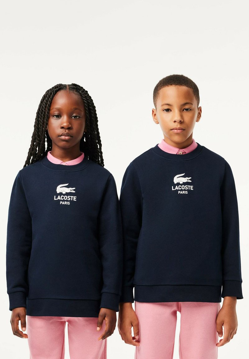 Sweatshirts bleu marine avec des poignets côtelés, arborant le logo blanc de Lacoste et le texte « PARIS » ; portés sur des chemises à col roses et des pantalons assortis.