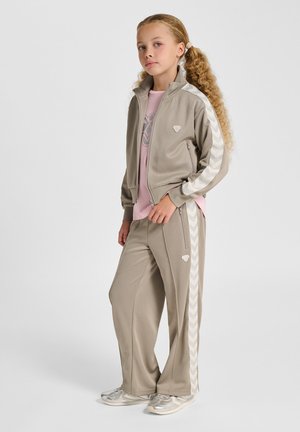 Beżowy dres z bluzą na zamek z paskami w kształcie litery V i dopasowanymi spodniami. W zestawie jasnoróżowa koszulka i srebrne sneakersy.