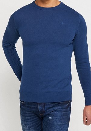 Mann trägt einen schlichten blauen Pullover mit Rundhalsausschnitt und langen Ärmeln sowie dunkelblaue Jeans, steht mit einer Hand am Rücken.