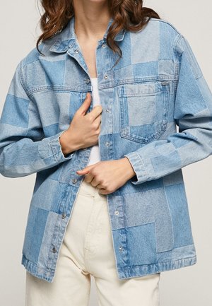 Chaqueta de mezclilla azul claro con un diseño de patchwork en diferentes tonalidades. Los botones a presión, un bolsillo en el pecho y detalles de costura añaden textura.