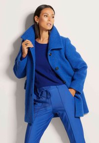 Blauwe wollen jas met grote kraag, sluiting met één knop en lange mouwen, gecombineerd met een marineblauwe trui en bijpassende blauwe broek.