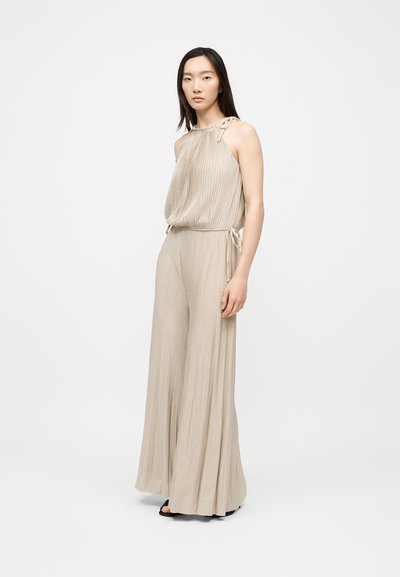 Femme aux cheveux raides et foncés portant un haut beige sans manches plissé avec épaule nouée et pantalon large assorti, debout sur fond blanc.
