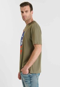 Hombre con camiseta gráfica verde oliva y jeans azules de perfil, con una mano en el bolsillo, contra un fondo claro y liso.