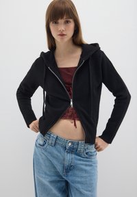 Felpa nera con zip e finitura testurizzata, dotata di cappuccio con coulisse e polsini a costine, indossata sopra una maglia bordeaux e jeans azzurri chiari.