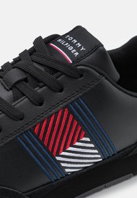 Sneaker in pelle nera con un design geometrico rosso e blu, dettagli testurizzati e chiusura con lacci. Branding Tommy Hilfiger sulla parte superiore.