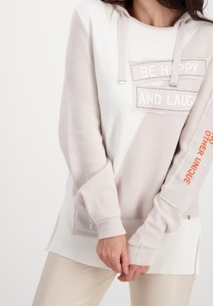 Kobieta ubrana w beżową i białą bluzę z kapturem z napisem "Bądź szczęśliwa i śmiej się" na piersi oraz "Bądź wyjątkowa" pomarańczowym kolorem na rękawie.