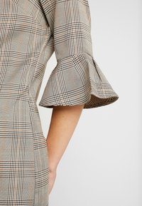 Rutigt blus med trumpetärmar. Tyget har en grå bas med svarta och orange rutmönster. Slät textur med en strukturerad passform.