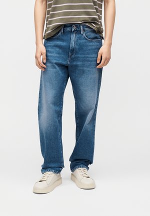 Personne portant un jean bleu droit, des baskets blanches et une chemise rayée verte et blanche rentrée, sur un fond uni.