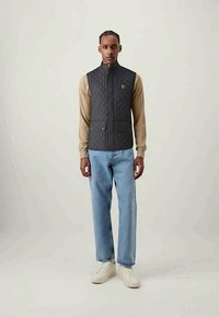 Vadderad mörkgrå väst med hög krage, framfickor och logotypdetalj, bärs över en beige långärmad tröja och ljusblå jeans.