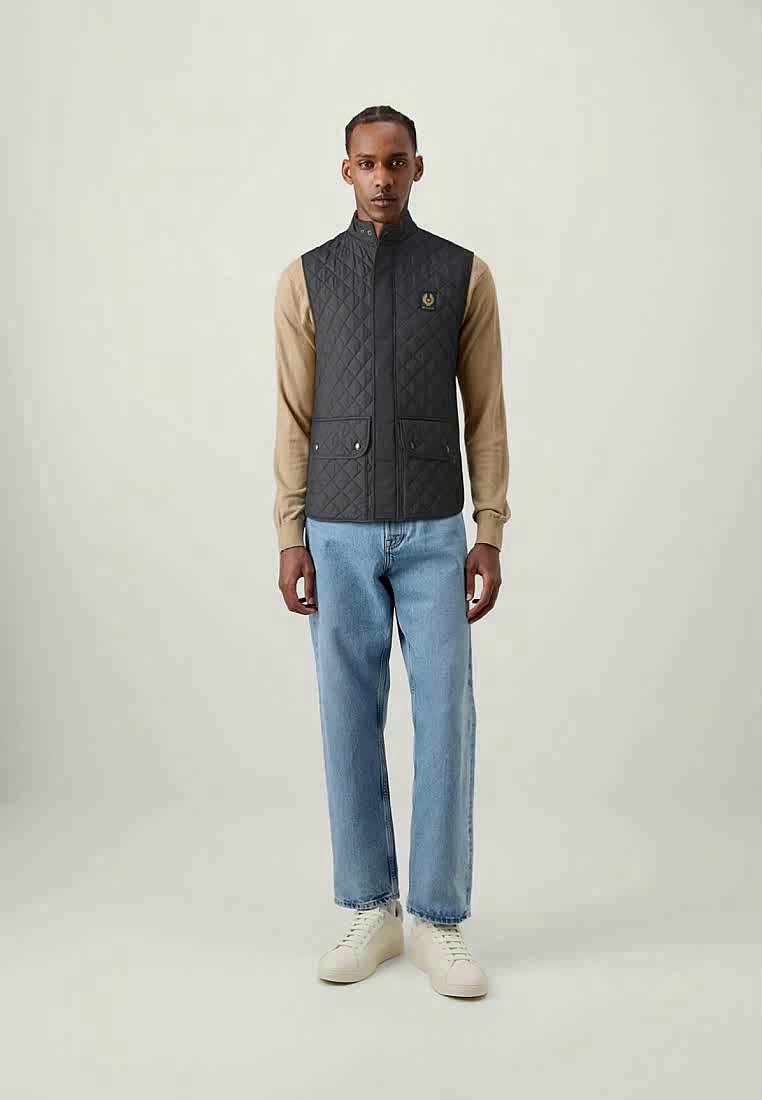 Gilet matelassé gris foncé avec col haut, poches avant et détail de logo, porté sur une chemise beige à manches longues et un jean bleu clair.