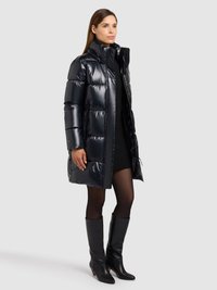 Čierna vatovaná bunda s vysokým golierom, lesklým povrchom a prešívaným dizajnom. Nosená cez tmavý sveter a doplnená čiernymi čižmami po kolená.