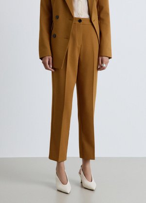 Persona che indossa un vestito marrone su misura con pantaloni corti e scarpe bianche con tacchi alti, in piedi davanti a uno sfondo uniforme.