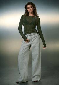 Maglia a maniche lunghe color oliva trasparente con accenti scintillanti, abbinata a jeans grigio chiaro a gamba larga. Tessuto liscio e vestibilità morbida, design minimalista.