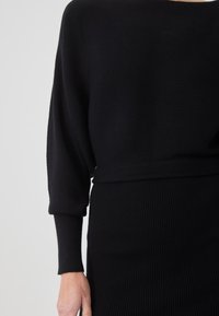 Top negru tricotat cu textură ribbed, având mâneci largi și talie ajustată. Materialul prezintă un model orizontal subtil.