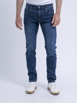 Homme portant un jean slim bleu foncé et des baskets blanches, debout sur un fond clair uni.