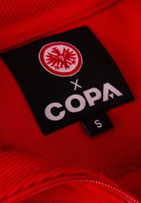 Rode hoodie stof met geribde kraag. Heeft een zwart label met een rode embleem en witte tekst "COPA," maatlabel toont "S."