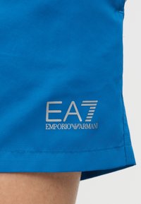 Μπλε υφασμάτινη βερμούδα με το λογότυπο "EA7 Emporio Armani" τυπωμένο σε λευκό κοντά στο τελείωμα πάνω από γυμνό πόδι.