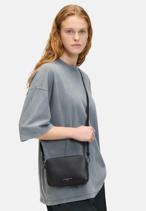 Borsa a tracolla nera in pelle con forma rettangolare, texture liscia e dettaglio del logo sulla parte anteriore; indossata con una t-shirt grigia oversize.