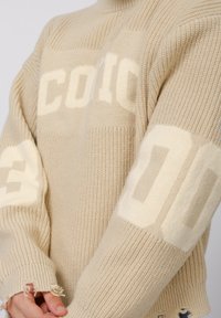 Beige knitted sweater with ribbed texture, featuring cream-colored block text "COCO" and "00" on sleeves, frayed cuffs and relaxed fit.