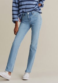 Jean en denim bleu clair à coupe slim, avec fermeture par boutons, cinq poches et des détails de couture subtils. Porté avec un haut rayé et des baskets blanches.