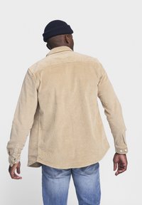 Chemise en velours côtelé beige avec un col boutonné, des manches longues et des fentes latérales, présentant un tissu texturé et un subtil détail de côtes.