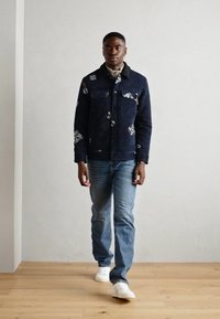 Μπλε υφασμάτινο μπουφάν με λουλουδάτα σχέδια, δύο μπροστινές τσέπες, φορεμένο πάνω από ανοιχτόχρωμο πουκάμισο. Συνδυασμένο με ανοιχτό μπλε τζιν και λευκά αθλητικά παπούτσια.