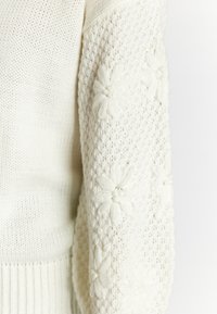 Weißer Strickpullover mit einem strukturierten Körper und strukturierten Ärmeln, verziert mit floralem Stickerei. Rippbündchen am Saum und an den Ärmeln sorgen für Stabilität.