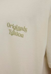 Kreminės spalvos marškinėliai su trumpomis rankovėmis, turintys iškeltą žalią tekstą "Originals Edition" priekyje. Minkšta, tekstūruota medžiaga.