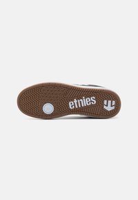 Talpă din cauciuc maro cu un model texturat, având branding alb "etnies". Partea superioară are un design alb-negru cu o finisare netedă.