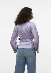 Veste en satin lilas, à manches longues, nouée à la taille. Elle présente une texture lisse et des accents en dentelle aux bords des manches. Portée avec du denim.
